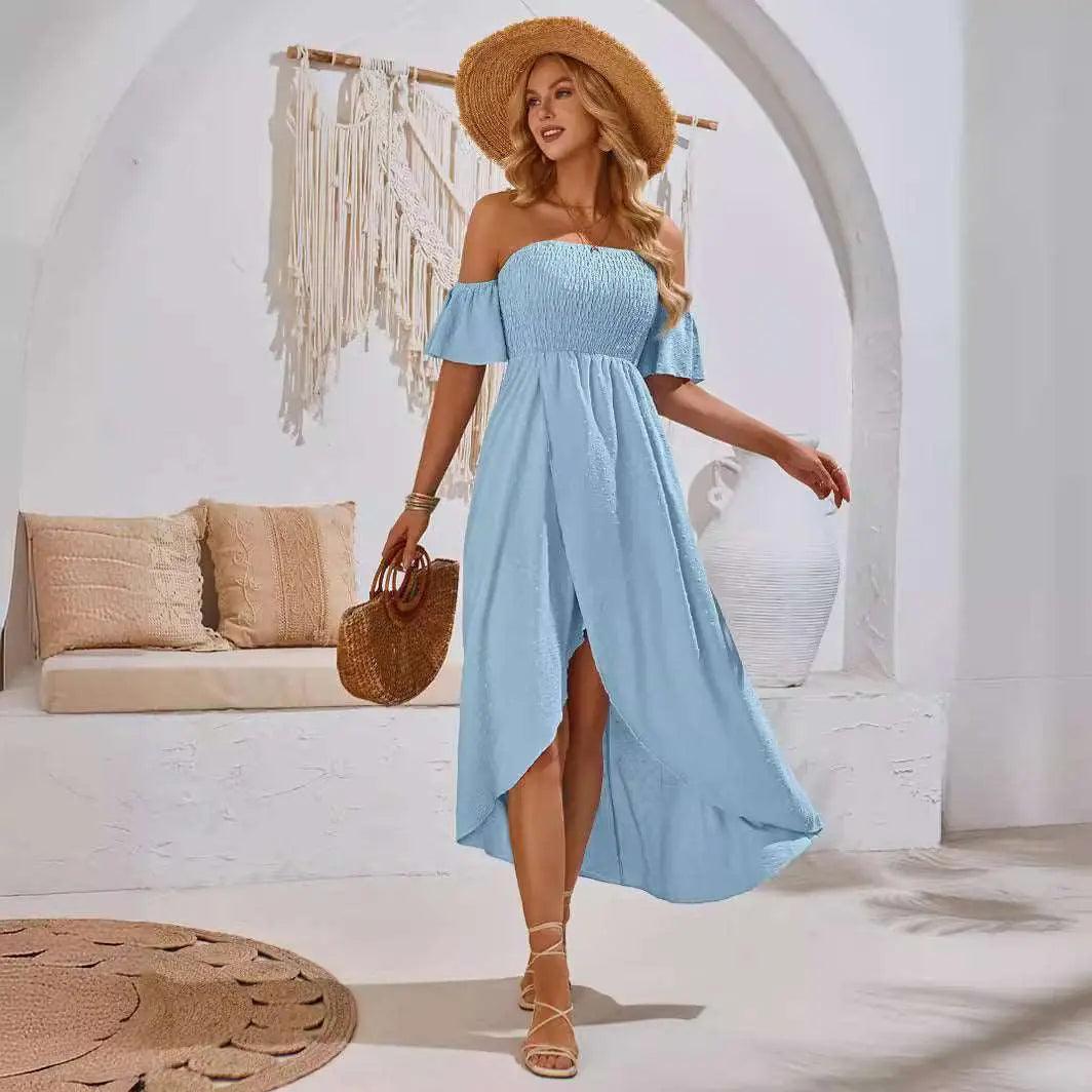 A woman wearing a blue, sleeveless, off-shoulder slit dress with a V-neckline, posing in a room with a white wall and a hat hanging in the background.