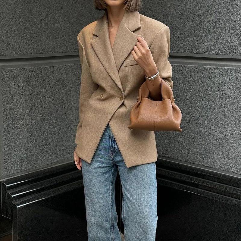 A woman wearing a beige business suit coat with long sleeves, paired with blue jeans, carrying a brown handbag, and standing against a grey background.