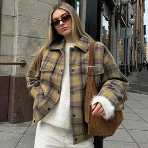 A woman wearing a plaid woolen jacket, sunglasses, and a brown purse, standing on a sidewalk.