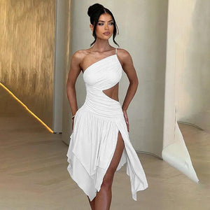 A woman wearing a white, solid color, form-fitting dress with a asymmetrical neckline and a high slit on the left side, standing in a room with a minimalist design.