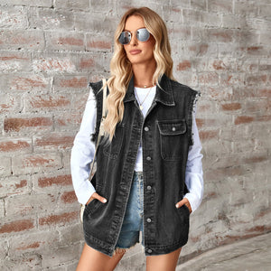 Woman wearing a black denim vest over a white shirt and denim shorts against a brick wall.