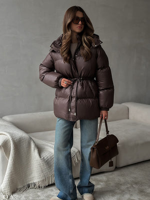 Woman wearing a brown puffer jacket and sunglasses, standing in a room with a gray wall and white sofa.