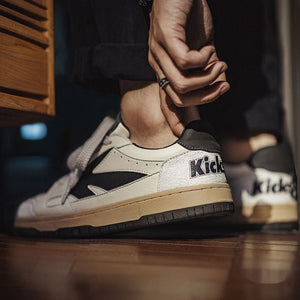 Close-up of a person wearing beige and black sneakers with 'Kickz' branding on a wooden floor.