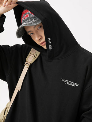 Person wearing a black hoodie with branding and a cap, standing against a neutral background.