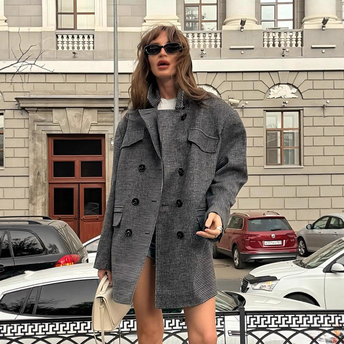 Woman in a gray coat standing on a street with cars and a building in the background