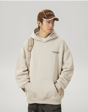 Person wearing a beige hoodie and brown cap with text, standing against a plain background.
