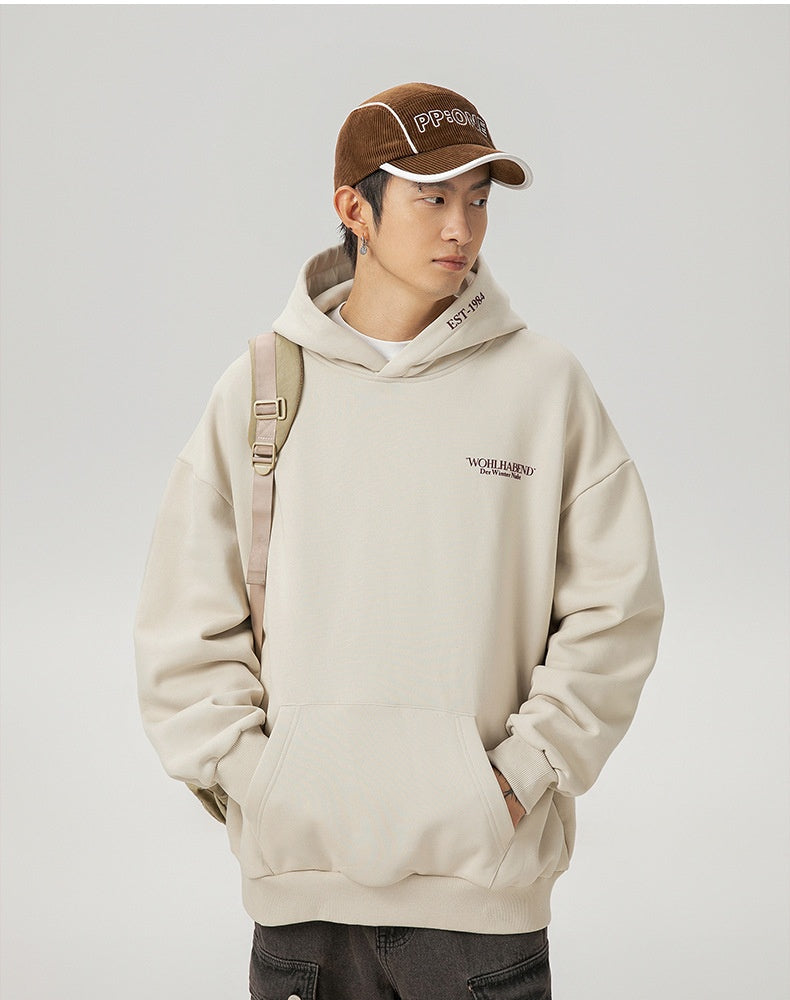 Person wearing a beige hoodie and brown cap with text, standing against a plain background.