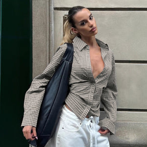 Woman wearing a checkered shirt and white pants, holding a black jacket against a neutral wall.