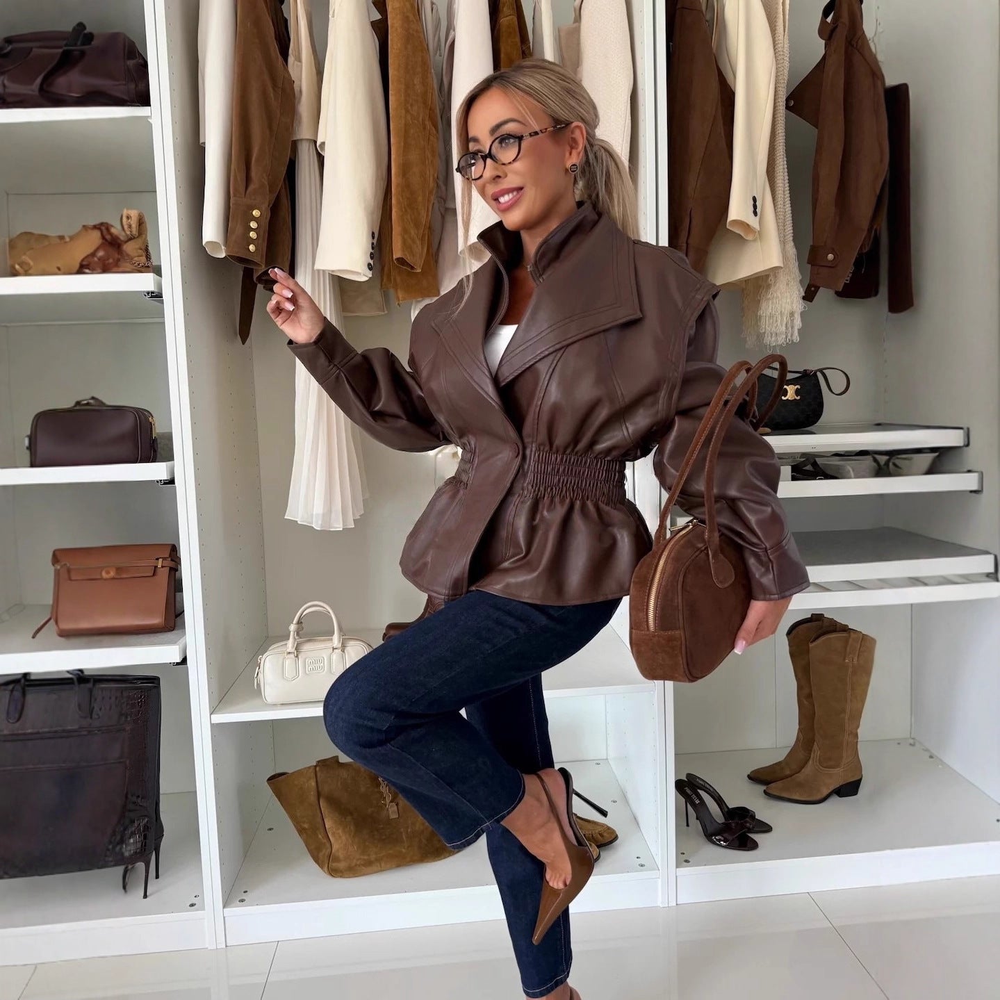 Woman in a brown leather jacket sitting in a closet with clothing and bags around her.
