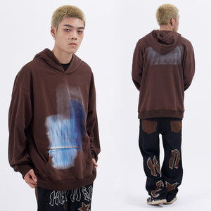 Person wearing a brown hoodie with a blue paint splatter design, standing in front of a white background.