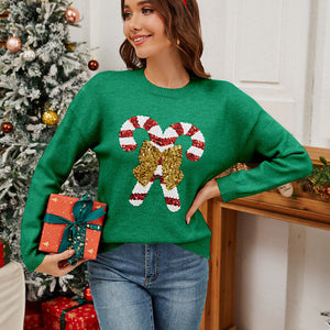 Woman wearing a green sweater with candy cane design, holding Christmas presents, in front of a decorated tree.