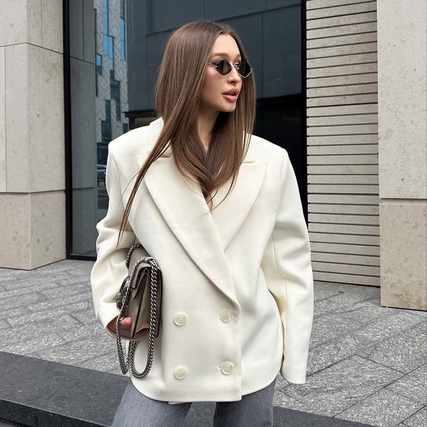 Woman wearing a white coat and sunglasses, standing on a city street.